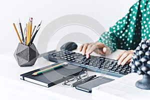 Close-up of hands middle-aged woman in green blouse typing on keyboard laptop computer, concrete holder with pencils and pens,