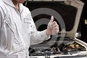 Close up hands of professional young mechanic man showing thumb up as sign of success with car in open hood at the garage backgrou