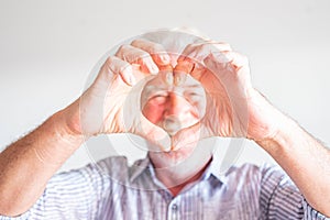 Close up of hands of senior or mature man doing a heart with his hands looking at the camera in the background - love and