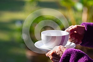 Close-up of hands senior woman`s holding a white coffee cup while standing in the garden. Space for text. Concept of aged people