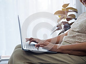 Close-up hands of senior woman working online, using laptop computer, sitting on the sofa at home.