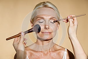 Close up of hands of skillful artist doing natural makeup for mature blonde woman  over beige background. The