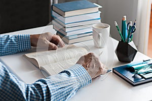 Close-up hands of the teacher man is preparing for the lesson: reading a book and making notes at the table, next to a pile of