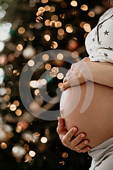 Close up of hands touching belly of young pregnant woman on a background of Christmas tree and lights. Expectation. Happy new year