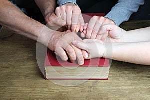 Close-up of the hands of two men and a woman folded in a prayer gesture on a thick family bible in a dark red cover, the concept