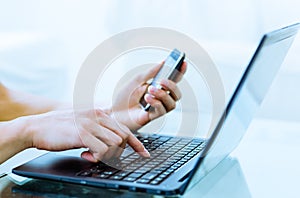 Close-up of hands typing on laptop computer while using a cell phone.