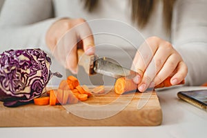 Close up on hands of unknown caucasian woman cutting carrot on the wooden board in the kitchen preparing vegan or vegetarian meal