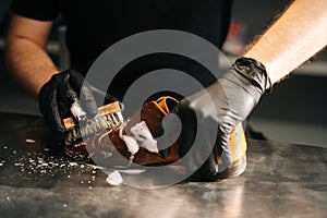 Close-up hands of unrecognizable shoemaker in black gloves applying cleaning foam on old light brown leather shoes.