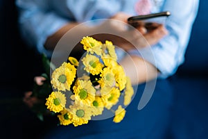 Close-up of hands of unrecognizable woman holding bouquet yellow chrysanthemums and using cell phone