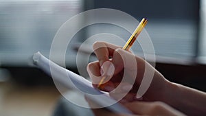 Close-up hands of unrecognizable woman making notes in paper notebook with pen sitting on chair on background of window