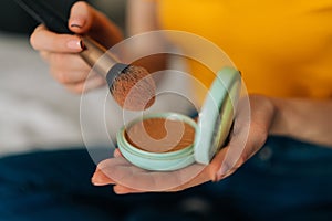 Close-up hands of unrecognizable young woman holding make up powder brush and case using it sitting on bed in bedroom