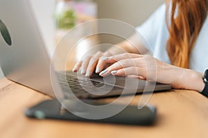 Close-up hands of unrecognizable young woman typing text on laptop keyboard sitting at table by window in cafe.