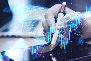 Close up of hands using digital pad on desktop with laptop and glowing candlestick forex chart map hologram on blurry background.