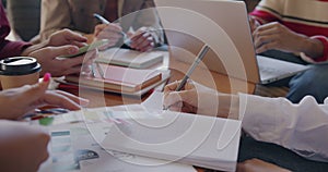 Close-up of hands using gadget and laptop writing in notebook while group of employees working in office at table