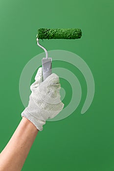 Close up of hands in working gloves holding middle-sized roller with green paint in front of green wall