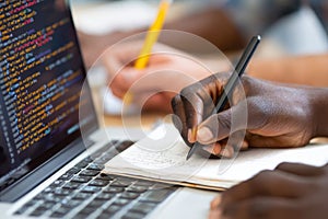 Close-up of hands writing notes while coding on a laptop in a collaborative workspace