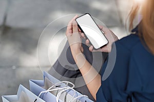 Close-up hands of young woman using empty smartphone while sitting in shopping mall area next to shopping bags.