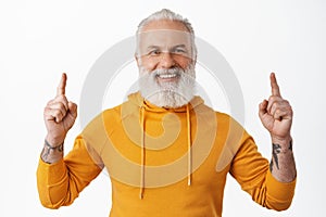 Close up of handsome hipster senior pointing fingers up and smiling happy. Old man with beard and tattoos showing