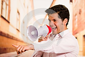 Close up of a handsome man screaming with a megaphone, pointing his hand to someone, in a blurred city background