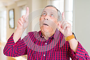 Close up of handsome senior man at home amazed and surprised looking up and pointing with fingers and raised arms