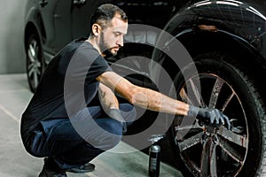 Close up of handsome young male employee worker cleaning the wheel rim of modern black crossover car by scrubbing brush