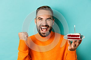 Close-up of happy adult man celebrating birthday, holding bday cake with candle and making wish, standing against