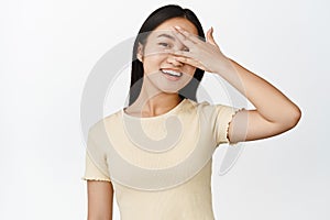 Close up of happy asian woman open eye, looking through fingers, smiling and laughing, white background