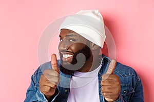Close-up of happy Black bearded guy in beanie showing support, agree or approve something, giggle devious and showing