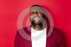 Close-up of happy and carefree Black man having fun, laughing and smiling, standing in blazer against red background