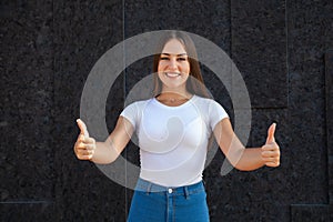 Close-up of a happy Caucasian girl showing thumbs up ahead, looking at the camera and dressed in a white T-shirt