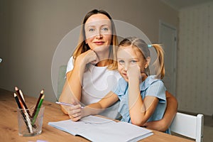 Close-up of happy elementary daughter doing homework with young mother sitting at home table by window, looking at