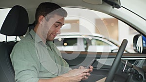 Close-up of happy handsome young man sitting behind wheel of luxury car and using mobile phone for surfing internet