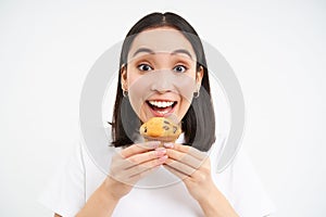 Close up of happy korean woman eating cupcake, delicious dessert, smiling from pleasure, white background