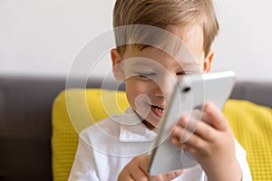 Close-up happy little cute preschool baby boy talking video conference by smartphone in living room at home on yellow gray sofa