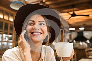 Close up of a happy lovely girl in hat sitting at the cafe table indoors, holding cup of tea,