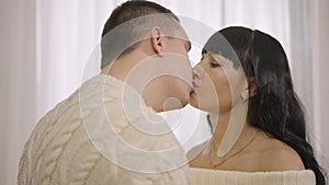 Close-up of happy loving Caucasian couple kissing in slow motion indoors. Portrait of beautiful woman and handsome man