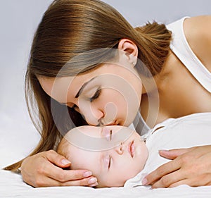 Close up of happy mother kissing her little sleeping baby lying on the bed at home