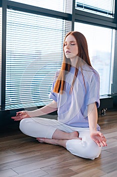 Close-up of happy relaxed young woman meditating sitting on floor in lotus pose on background of window in light office