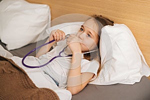 Close-up of happy sick little girl playing with stethoscope while lying in bed in bedroom during visit of pediatrician