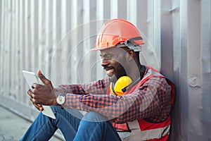 Close up happy smiling male African American engineer wearing safety vest and helmet, sitting and using mobile phone at logistic