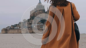 Close-up happy tourist woman in elegant coat walking towards stunning Mont Saint Michel castle in Normandy slow motion.