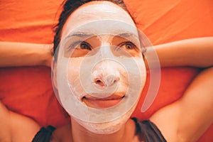 Close up of happy young beautiful smiling woman with white facial clay mask relaxing on the bed