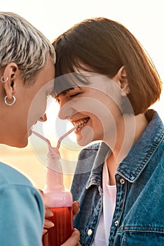 Close up of happy young lesbian couple having fun, drinking from one glass bottle with the straw, Two women enjoying