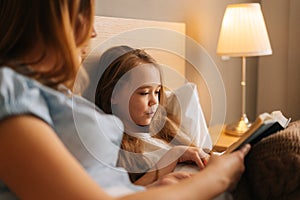 Close-up of happy young mother and adorable daughter reading together children book before going to sleep while lying in
