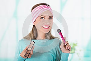 Close up of happy young woman holding a make up palette and doing crazy make-up in her face using a make up brush, in a