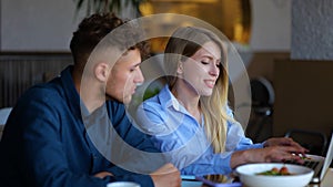 Close up of happy young woman sitting at table in restaurant talking with handsome man and typing on a laptop. Caucasian