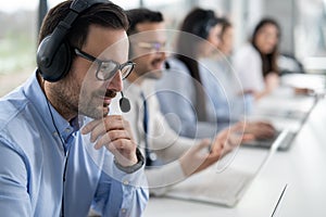 Close up head shot of handsome male customer support agent with headset working in call center. Group of helpdesk operators at photo