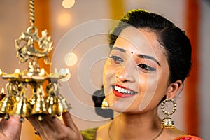 close up head shot of happy young indian girl lighting up hanging oil lamp during festival at home - concept of indian