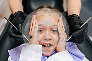 Close-up headshot portrait of cute afraid child girl holding head from fear with hand, sitting on dentist chair. Closeup