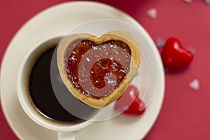 Close up heart shape cookie with strawberry jam. Cup of coffee, morning drink. Red background with copy space